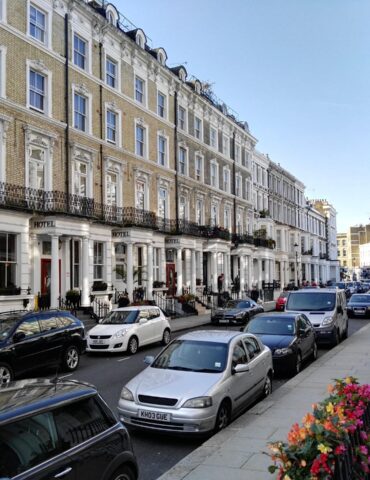 Row of classic Georgian townhouses with ornate window frames and black iron balconies, parked cars lining a sunny street. Flowers along the sidewalk railings.