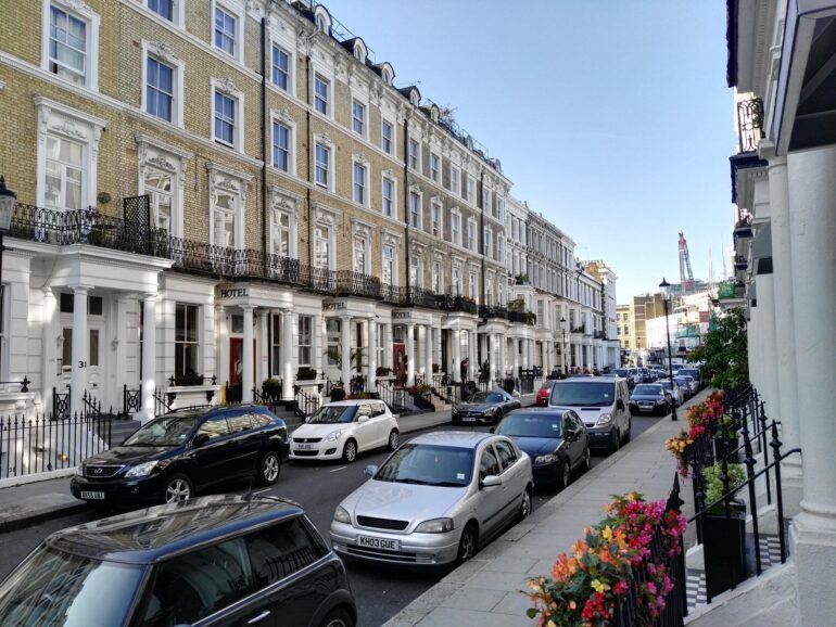 Row of classic Georgian townhouses with ornate window frames and black iron balconies, parked cars lining a sunny street. Flowers along the sidewalk railings.