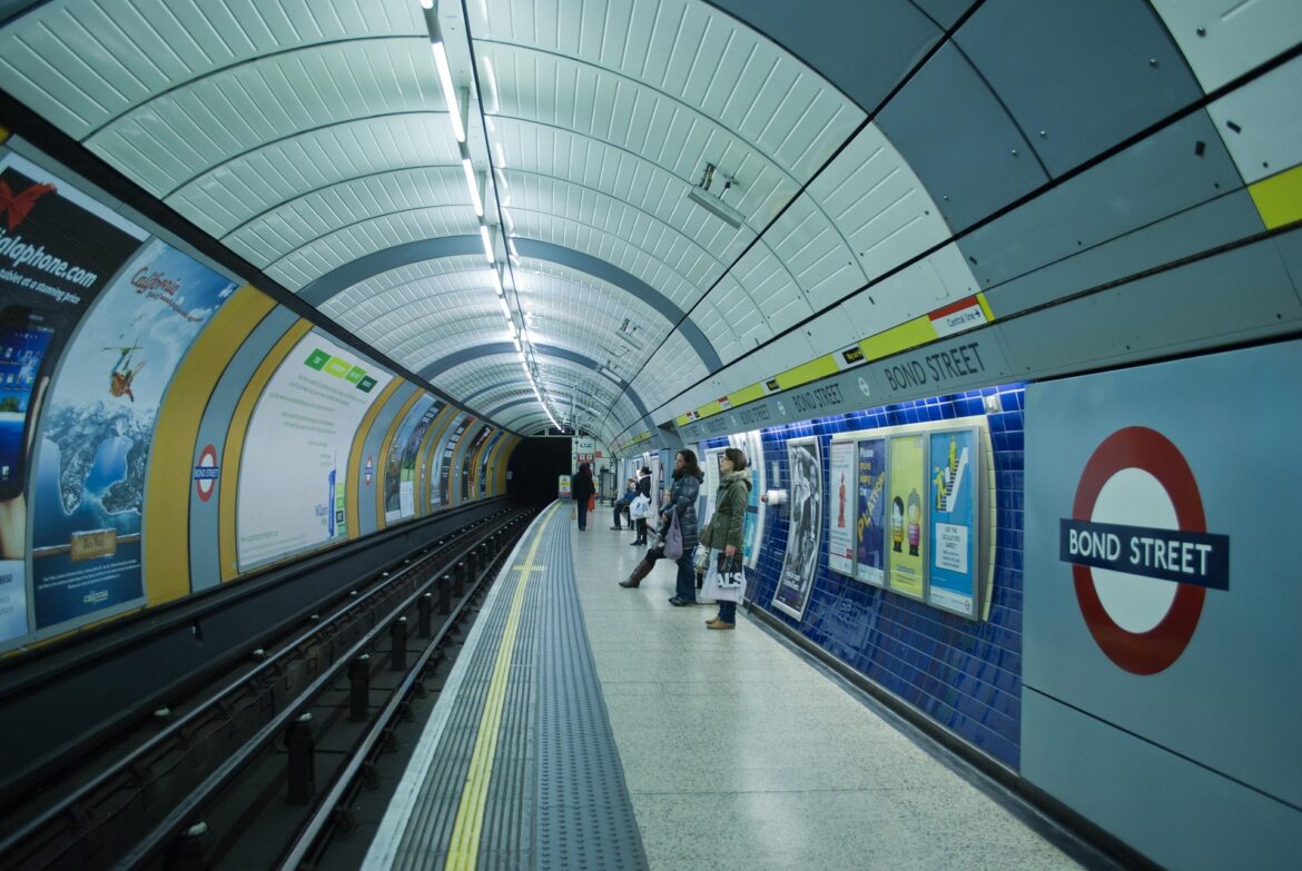 London Underground Bond Street platform with passengers waiting beside ads and curved ceiling panels.