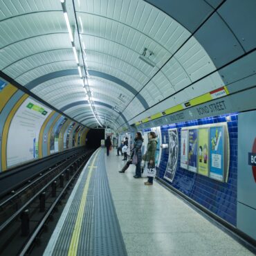 London Underground Bond Street platform with passengers waiting beside ads and curved ceiling panels.