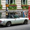 Vintage pale-blue Triumph Stag convertible parked on a city street beside a red British telephone booth; a couple walks by.