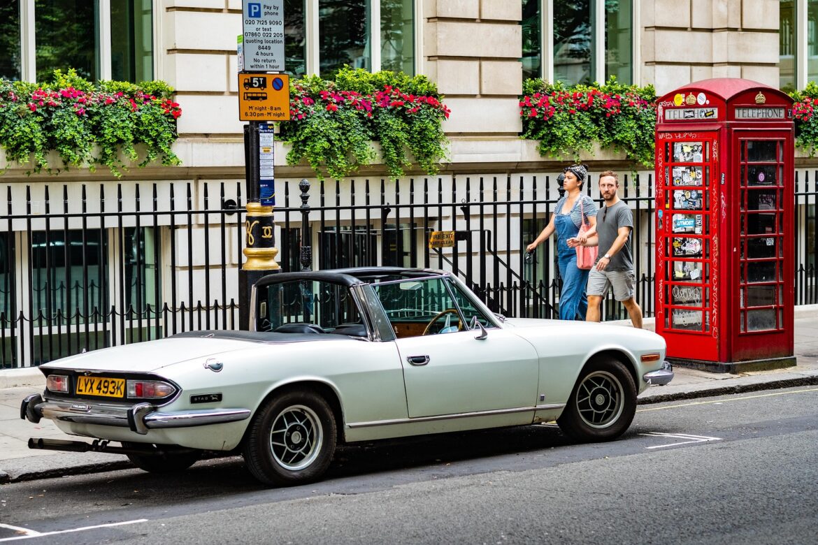 Vintage pale-blue Triumph Stag convertible parked on a city street beside a red British telephone booth; a couple walks by.