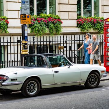 Vintage pale-blue Triumph Stag convertible parked on a city street beside a red British telephone booth; a couple walks by.