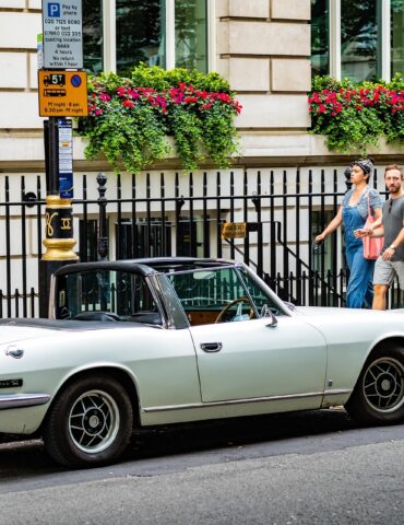 Vintage pale-blue Triumph Stag convertible parked on a city street beside a red British telephone booth; a couple walks by.