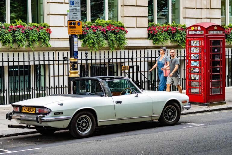 Vintage pale-blue Triumph Stag convertible parked on a city street beside a red British telephone booth; a couple walks by.