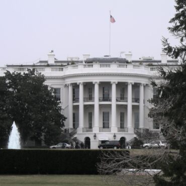 White neoclassical building with tall columns and an American flag on the roof, viewed behind a fountain and trees on the lawn.