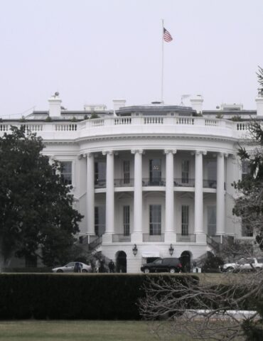 White neoclassical building with tall columns and an American flag on the roof, viewed behind a fountain and trees on the lawn.