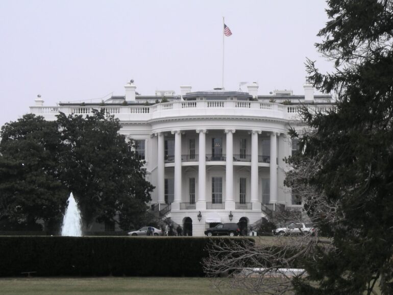 White neoclassical building with tall columns and an American flag on the roof, viewed behind a fountain and trees on the lawn.