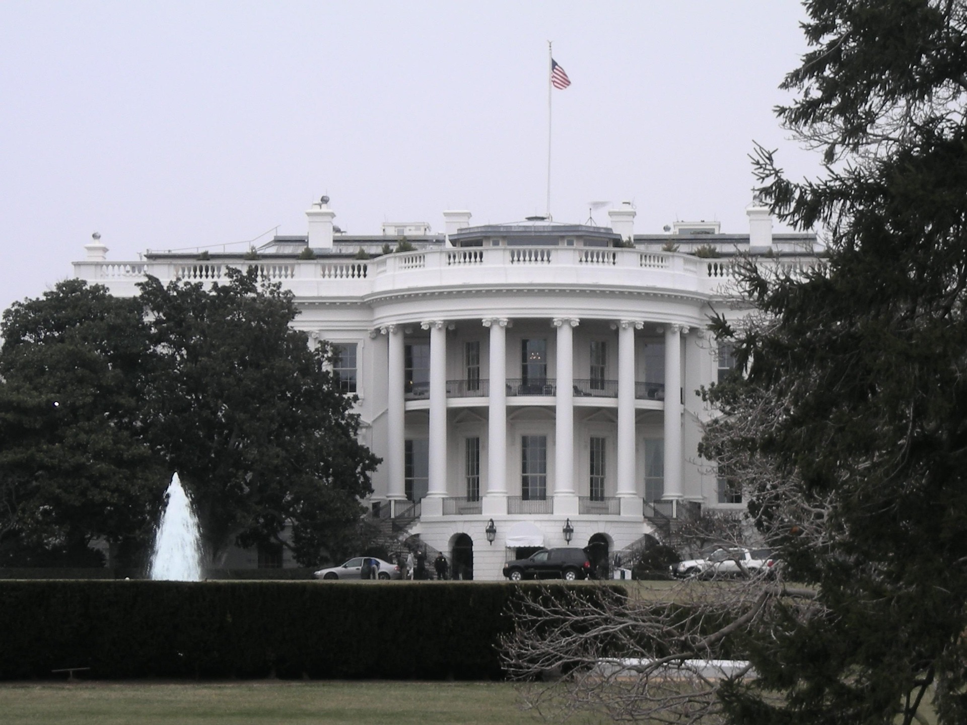 White neoclassical building with tall columns and an American flag on the roof, viewed behind a fountain and trees on the lawn.