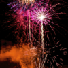 Fireworks explode over a medieval castle wall at night, with silhouetted spectators watching from below.