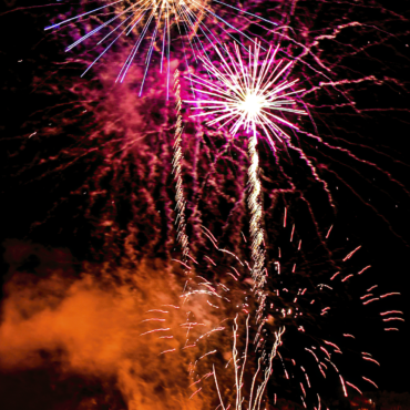 Fireworks explode over a medieval castle wall at night, with silhouetted spectators watching from below.