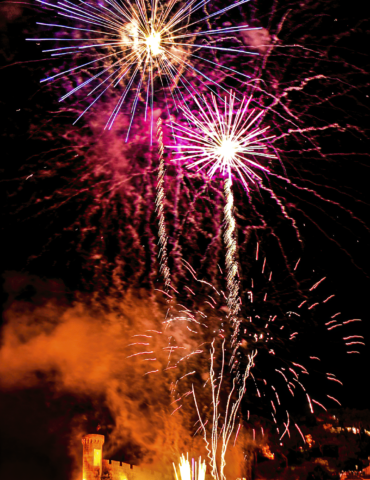 Fireworks explode over a medieval castle wall at night, with silhouetted spectators watching from below.