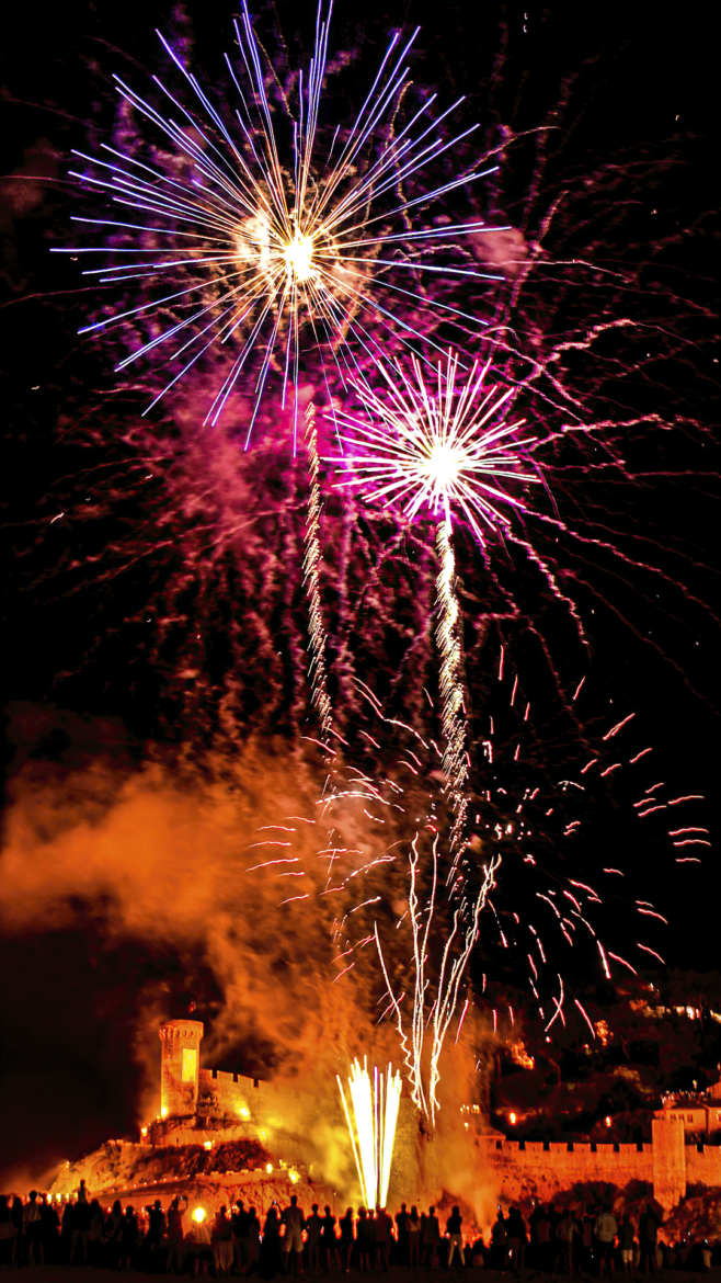 Fireworks explode over a medieval castle wall at night, with silhouetted spectators watching from below.