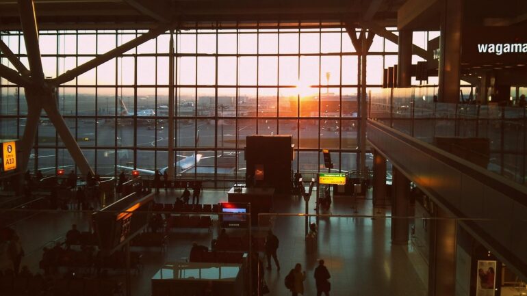 Sunset light pours through large glass walls of an airport terminal, with travelers seated and walking below.