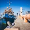 Blue fishing boat moored at a pier under a bright blue sky, with orange lifebuoys on the deck and ropes along the railing.