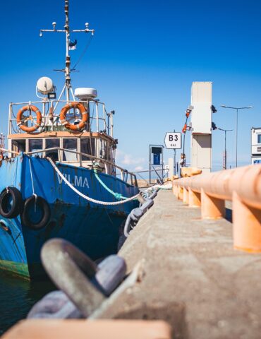 Blue fishing boat moored at a pier under a bright blue sky, with orange lifebuoys on the deck and ropes along the railing.