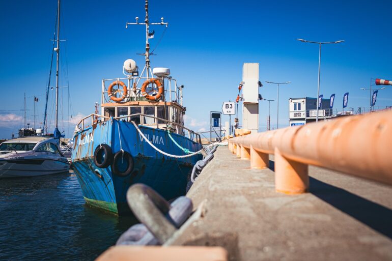 Blue fishing boat moored at a pier under a bright blue sky, with orange lifebuoys on the deck and ropes along the railing.
