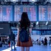 Woman with a black backpack standing in an airport terminal, facing large flight information boards above the concourse.