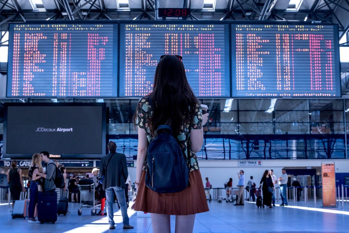 Woman with a black backpack standing in an airport terminal, facing large flight information boards above the concourse.
