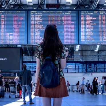 Woman with a black backpack standing in an airport terminal, facing large flight information boards above the concourse.