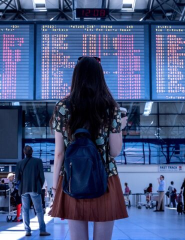 Woman with a black backpack standing in an airport terminal, facing large flight information boards above the concourse.