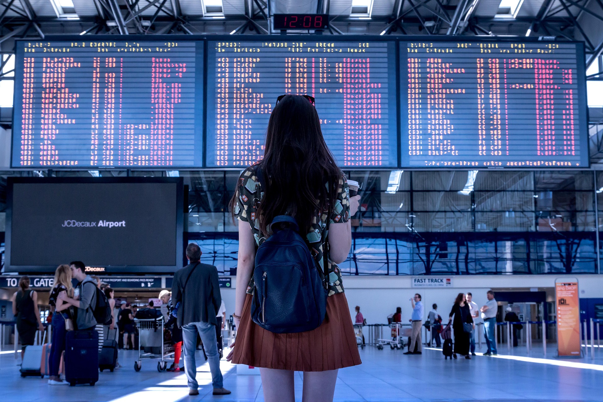 Woman with a black backpack standing in an airport terminal, facing large flight information boards above the concourse.