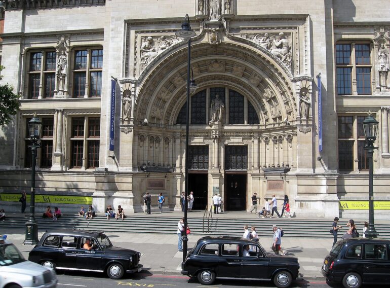 jorisamonen-building-255445_1920 - NOTE.radio - Polskie radio w UK | Polskie radio w Londynie Grand stone museum facade with an ornate arched entry, sculpted figures, and banner flags; people sit and walk on the steps while black taxis pass by on the street.