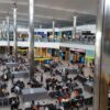 Busy airport departures hall with seating, shops, and two levels connected by glass railings and tall metallic columns.