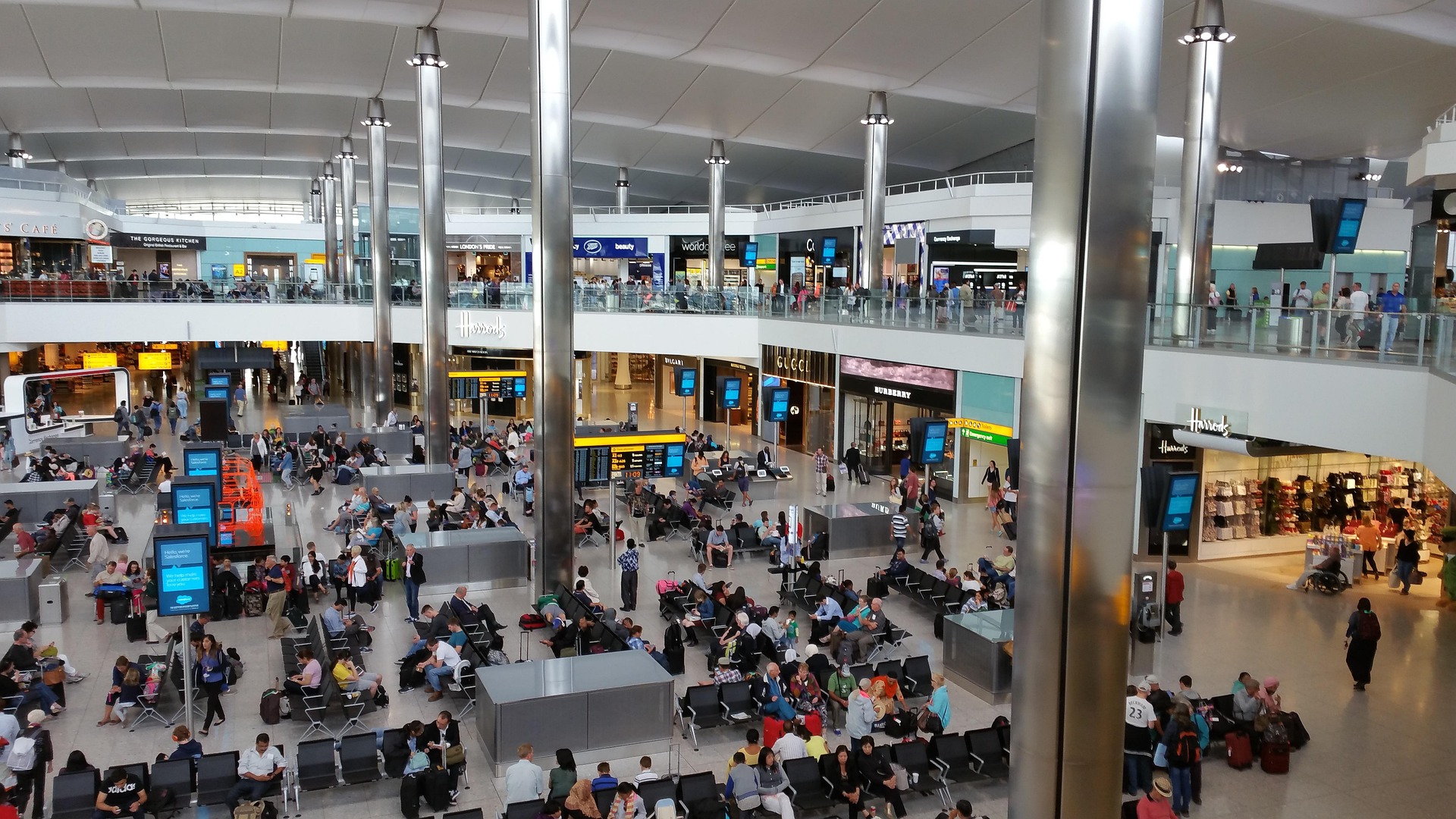 Busy airport departures hall with seating, shops, and two levels connected by glass railings and tall metallic columns.