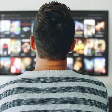 Person wearing a striped sweater sits facing a large wall-mounted TV showing a grid of streaming movie thumbnails (back view).