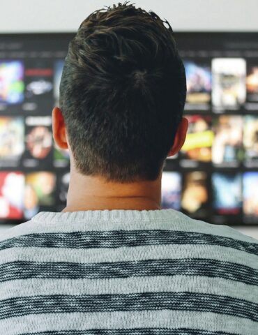 Person wearing a striped sweater sits facing a large wall-mounted TV showing a grid of streaming movie thumbnails (back view).