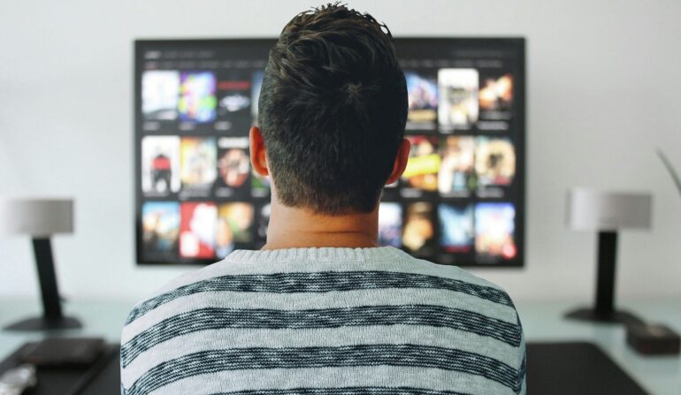 TV - NOTE.radio - Polskie radio w UK | Polskie radio w Londynie Person wearing a striped sweater sits facing a large wall-mounted TV showing a grid of streaming movie thumbnails (back view).