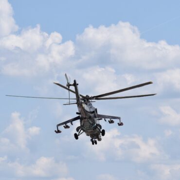 Military helicopter in flight with spinning rotor blades against a blue sky with clouds.