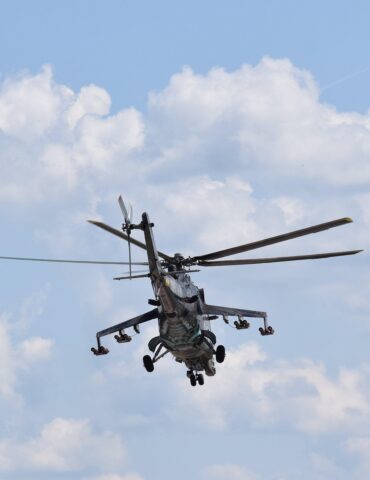 Military helicopter in flight with spinning rotor blades against a blue sky with clouds.