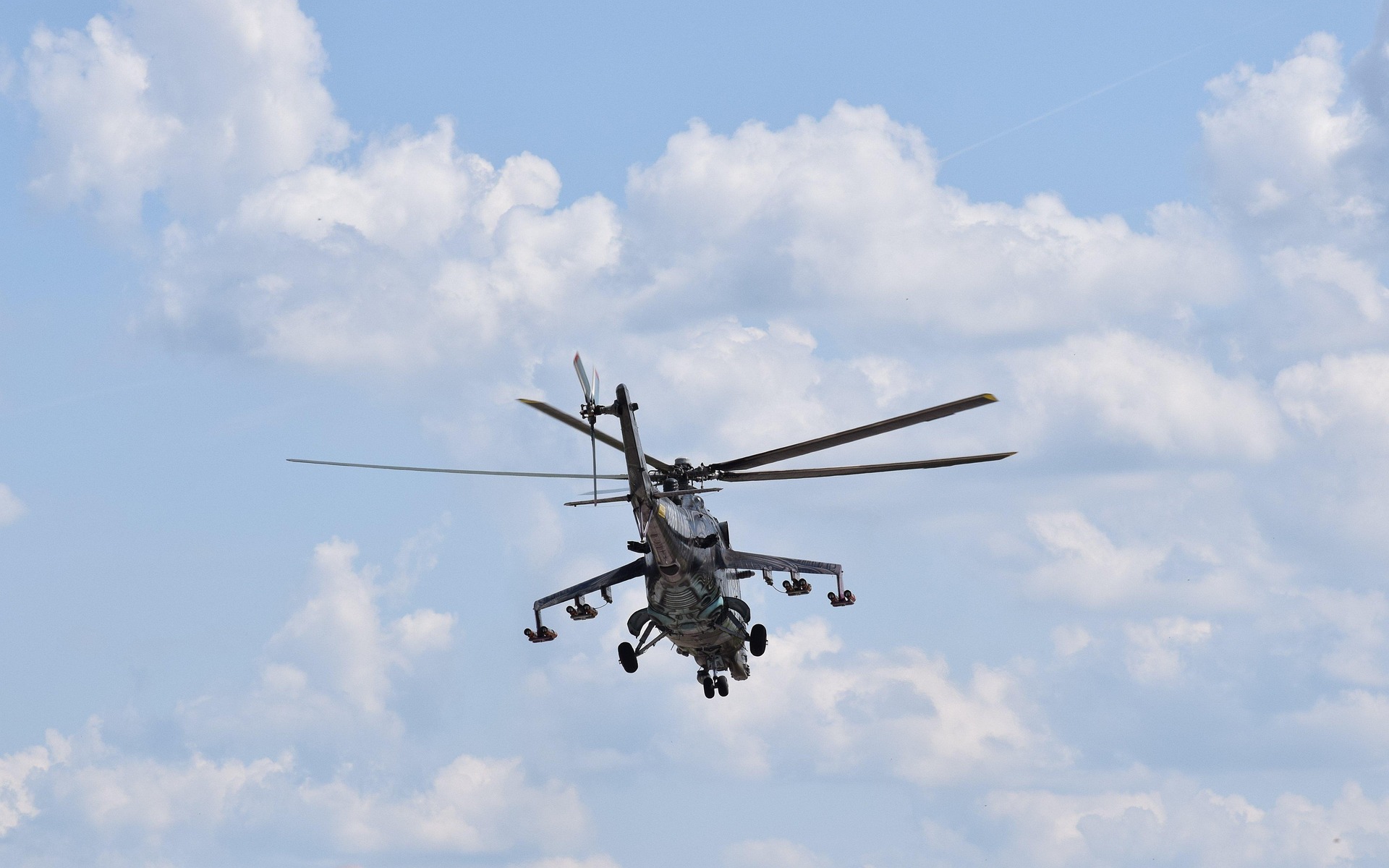 Military helicopter in flight with spinning rotor blades against a blue sky with clouds.