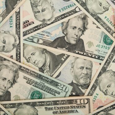 Overlapping U.S. dollar bills (various denominations) scattered in a collage scene, showing portraits and security features.