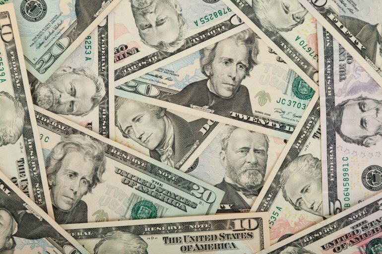 Overlapping U.S. dollar bills (various denominations) scattered in a collage scene, showing portraits and security features.