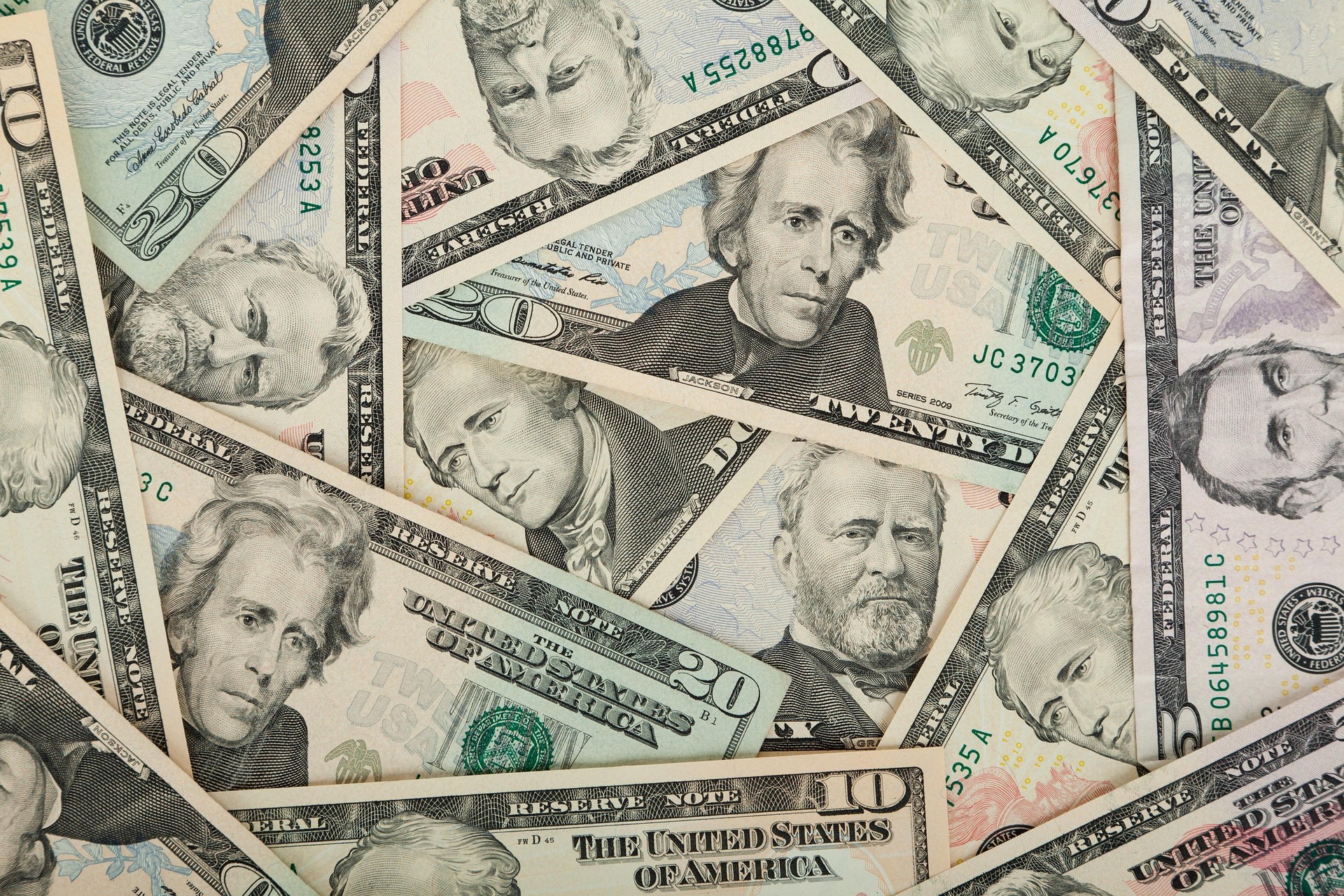 Overlapping U.S. dollar bills (various denominations) scattered in a collage scene, showing portraits and security features.