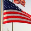 American flag waving on a flagpole against a clear blue sky, with another flag visible in the background