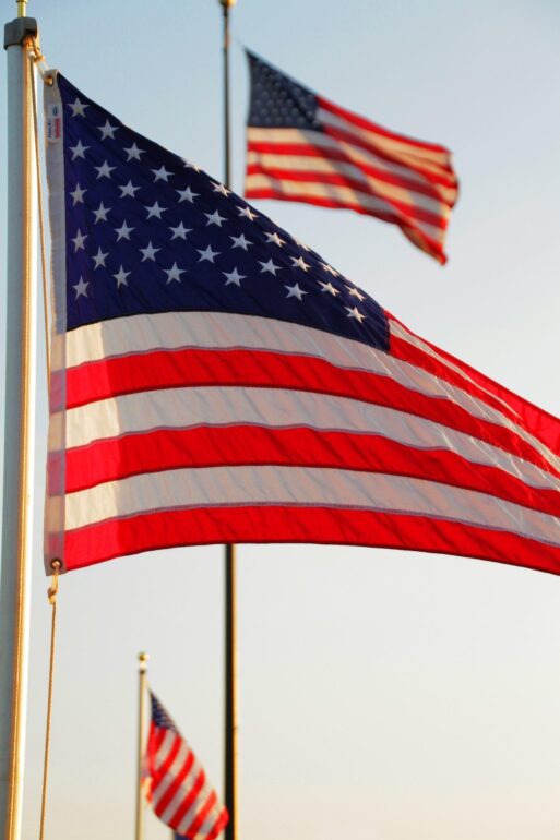 American flag waving on a flagpole against a clear blue sky, with another flag visible in the background
