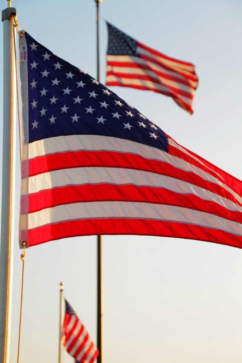 American flag waving on a flagpole against a clear blue sky, with another flag visible in the background