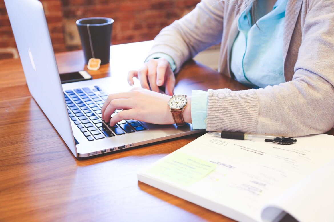 Person typing on a laptop at a wooden desk with a notebook and pen nearby.