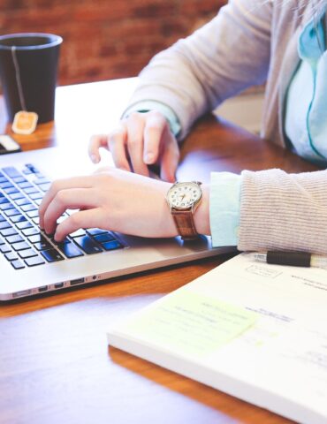 Person typing on a laptop at a wooden desk with a notebook and pen nearby.
