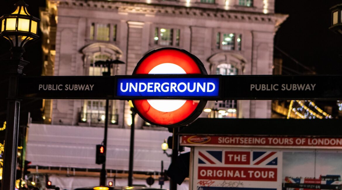 London Underground roundel sign at night, with blue 'Underground' banner and 'Public Subway' text on both sides against a city street backdrop