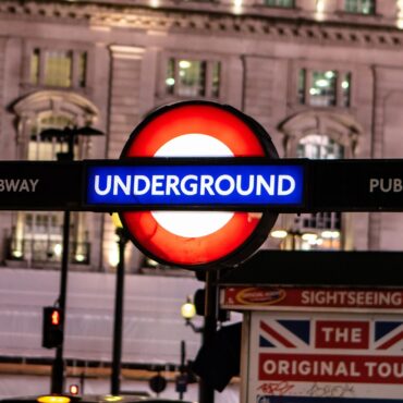 London Underground roundel sign at night, with blue 'Underground' banner and 'Public Subway' text on both sides against a city street backdrop