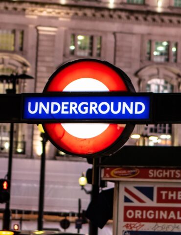 London Underground roundel sign at night, with blue 'Underground' banner and 'Public Subway' text on both sides against a city street backdrop