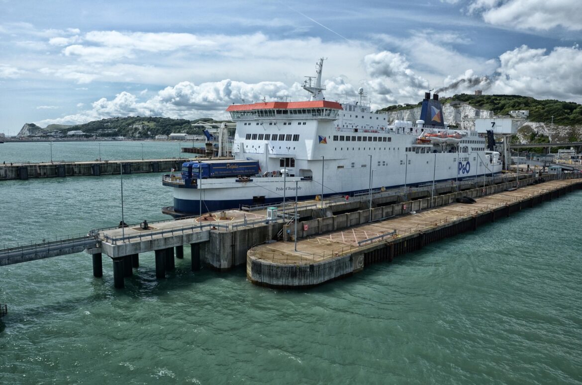Large white passenger ferry docked at a concrete pier with hills and blue sky in the background