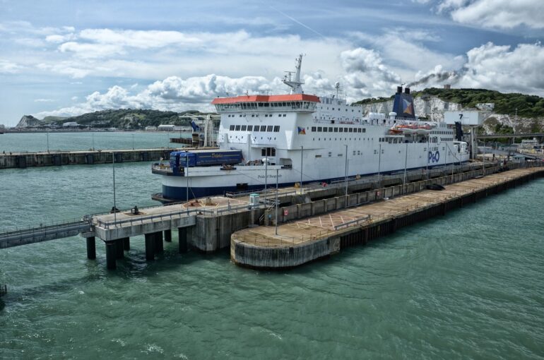Large white passenger ferry docked at a concrete pier with hills and blue sky in the background
