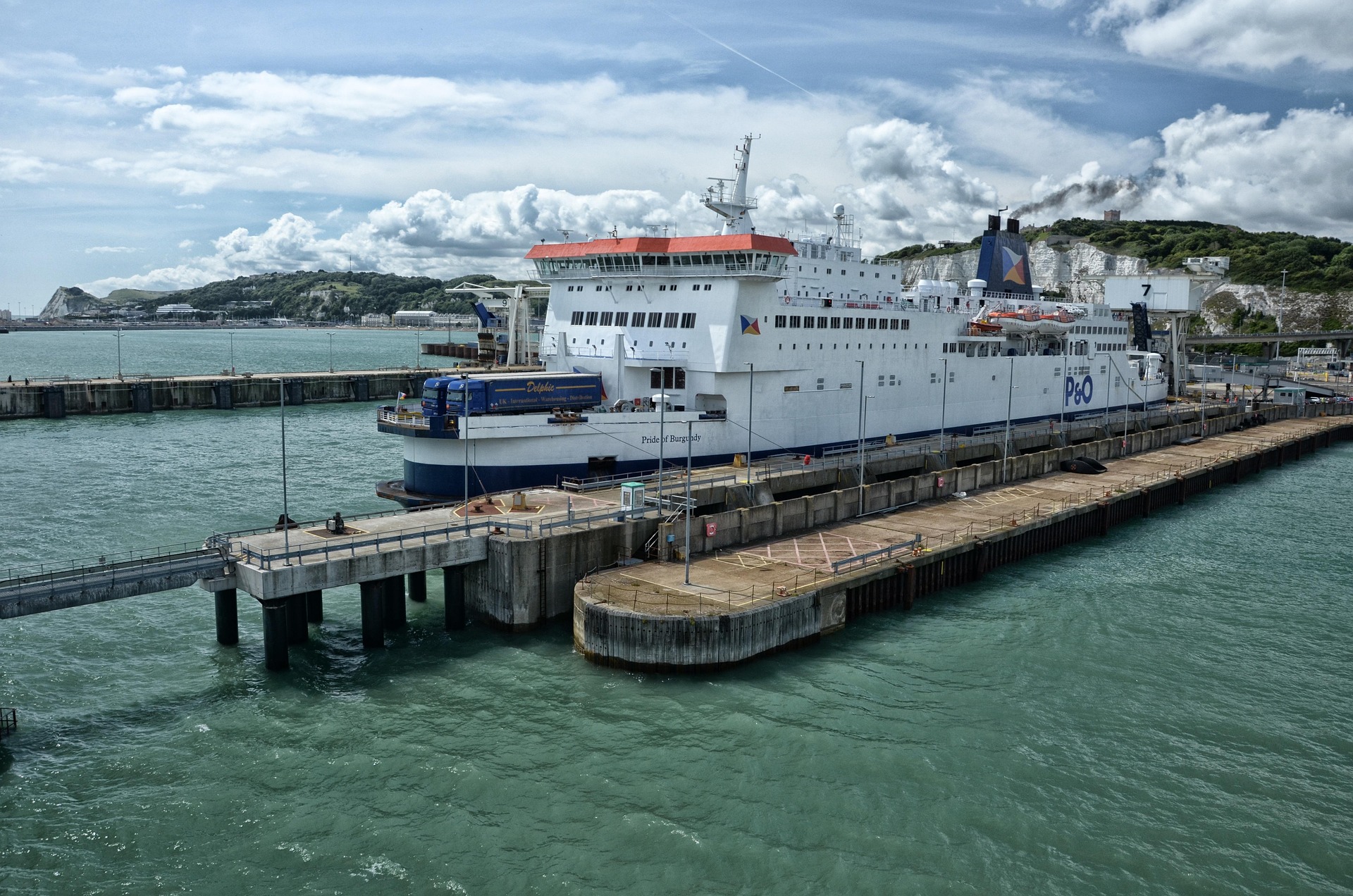Large white passenger ferry docked at a concrete pier with hills and blue sky in the background
