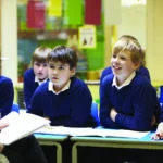 Teacher leads a small class discussion with students in blue sweaters seated at desks, notebooks open.